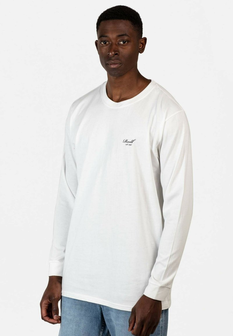 Langärmliges weißes T-Shirt aus Baumwolle mit Rundhalsausschnitt und einem kleinen schwarzen Logo auf der linken Brustseite. Getragen mit blauen Jeans.