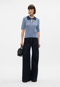 Ανοιχτό μπλε πλεκτό τοπ με σκούρο μπλε γιακά και λεπτομέρεια σε σχήμα κλειδαρότρυπας, συνδυασμένο με φαρδιά τζιν παντελόνια σε σκούρο denim και μια μικρή μαύρη πλεκτή τσάντα.