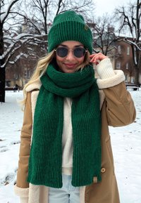 Grøn strikket hue og matchende tørklæde med ribbet struktur. Modellen bærer en cremefarvet sweater og en beige frakke med sælskinddetaljer i en sneklædt omgivelser.