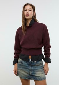Donna con lunghi capelli castani che indossa un maglione marrone bordeaux sopra una camicia nera a righe sottili, infilata in una minigonna di jeans sbiadita, in piedi contro uno sfondo semplice.