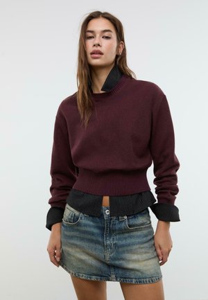 Donna con lunghi capelli castani che indossa un maglione marrone bordeaux sopra una camicia nera a righe sottili, infilata in una minigonna di jeans sbiadita, in piedi contro uno sfondo semplice.