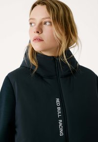Schwarze Daunenjacke mit hohem Kragen, ausgestattet mit einem Reißverschluss und weißem "RED BULL RACING"-Text auf der Vorderseite. Glatte, elegante Textur.