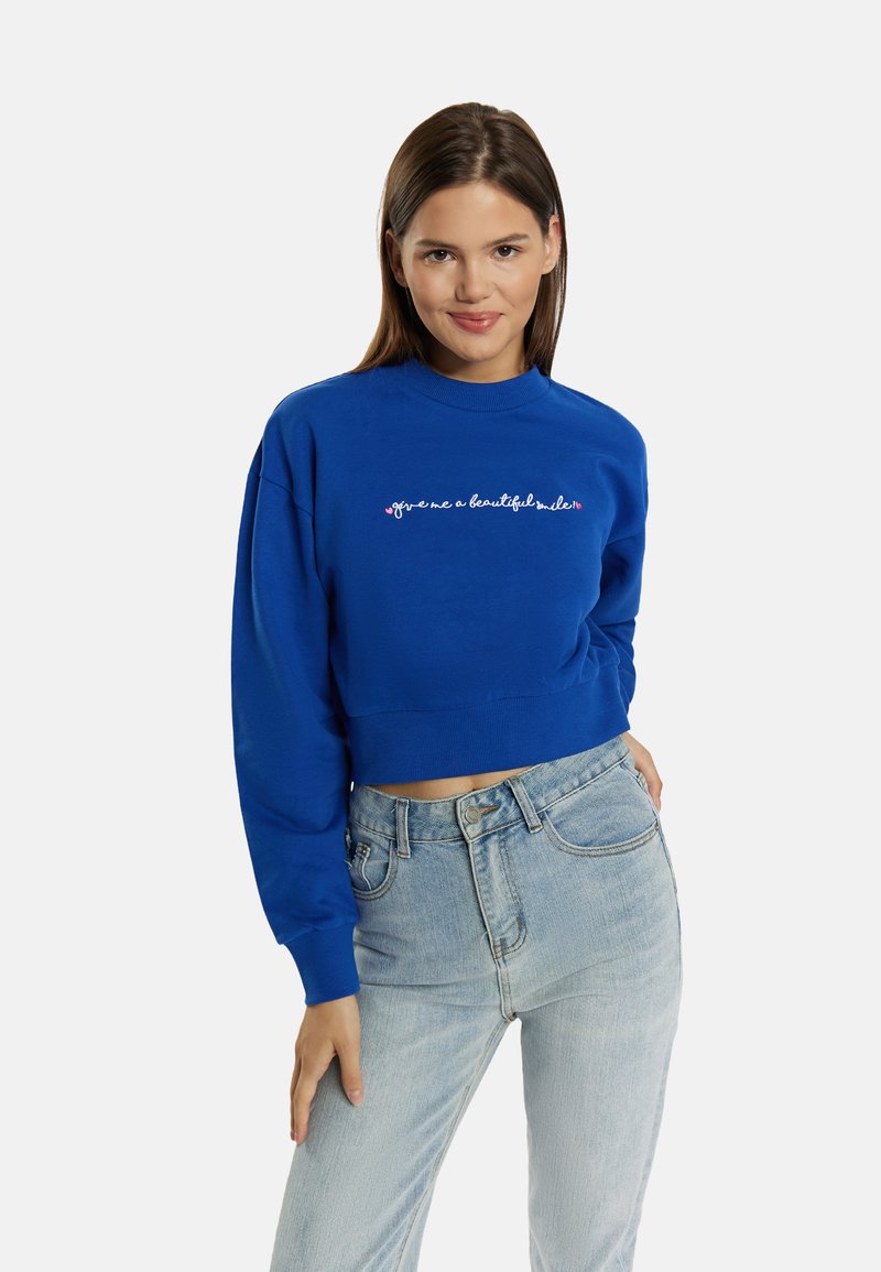 Felpa blu tagliata con maniche lunghe, con testo ricamato bianco e rosa sul petto. Abbinata a jeans azzurri chiari.