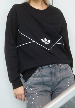 Osoba nosząca czarną bluzę Adidas z białymi lamówkami i logo, w połączeniu z szarymi jeansami z białymi plamami farby.
