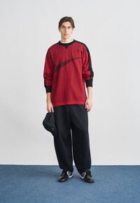 Červený a čierny svetlý sveter s vertikálnymi pruhmi a voľným strihom, doplnený voľnými čiernymi nohavicami a čiernymi teniskami; čiernu šiltovku drží v ruke.