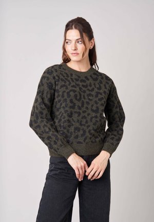 Femme portant un pull imprimé léopard vert foncé et noir à manches longues et un pantalon noir, regardant légèrement sur le côté.