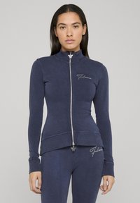 Marineblå glidelåsjakke i mykt materiale, med høy krage, tettsittende design og dekorert logo på brystet, kombinert med matchende bukser.