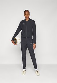 Crni Nike trenirku s jaknom na zip i hlačama s elastičnom paskom, s bijelim bočnim prugama i teksturiranim nogometnim loptom.