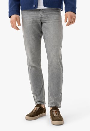 Muž oblečený v svetlosivých džínsoch, hnedých semišových teniskách s béžovými podrážkami a námorníckej modrej bunde stojaci na bielom pozadí.