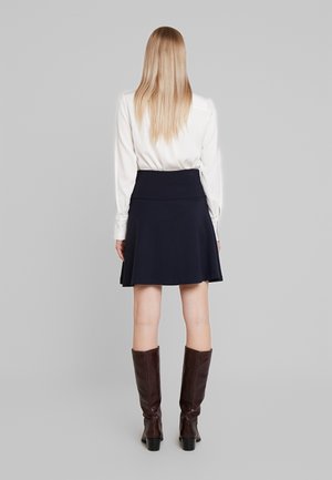 Vrouw met lang blond haar, gekleed in een witte blouse, een marineblauwe rok tot op de knie en bruine laarzen tot over de knie, staand met de rug naar de kijker op een grijze achtergrond.