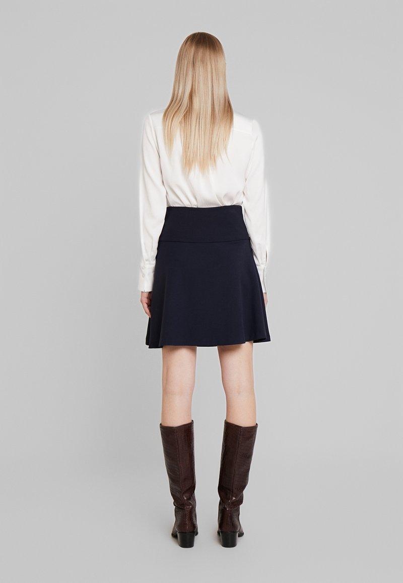Femme aux cheveux longs blonds portant un chemisier blanc, une jupe bleu marine arrivant aux genoux et des bottes marron montant jusqu'aux genoux, debout de dos sur un fond gris.