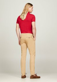 Polo shirt rossa con maniche corte, abbinata a pantaloni chinos beige. Scarpe marroni con lacci, leggero lucido sul tessuto e dettagli nella tasca posteriore.