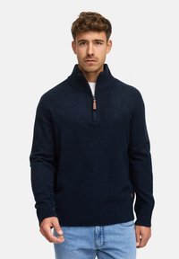 Marineblå zip-sweater i et blødt strikket stof med høj krave, ribbede ærmer og kant, med en lille læderdetalje nær kanten.