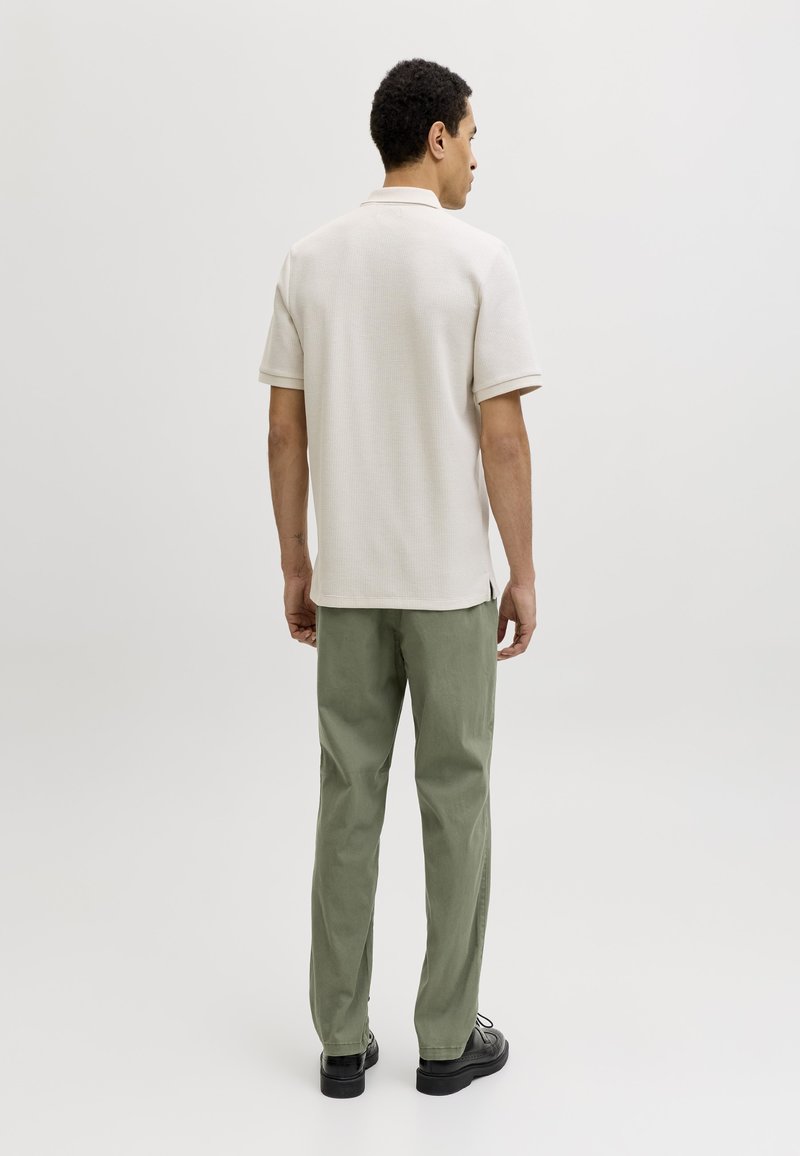 Hombre de pie de espaldas, vestido con una camisa polo beige de manga corta, pantalones verde claro y zapatos negros sobre un fondo claro y sencillo.