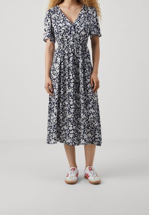 Kvinde iført en navyblå og hvid blomsterprintet midikjole med korte ærmer og hvide sneakers med røde striber, stående på en ensfarvet lys baggrund.
