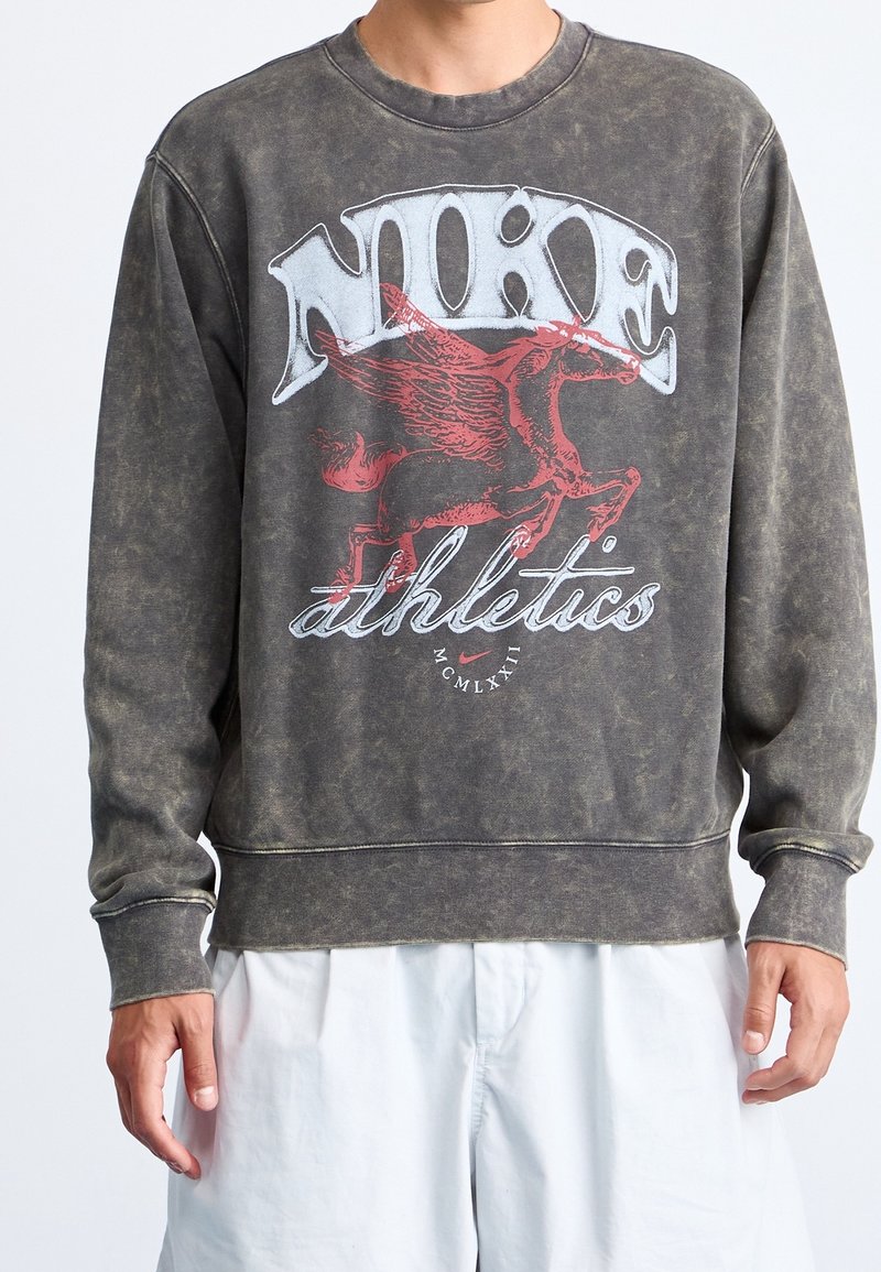 Personne portant un sweat-shirt noir délavé avec le texte « NIKE athletics » et un graphisme rouge de cheval ailé sur le devant, associé à un short blanc.