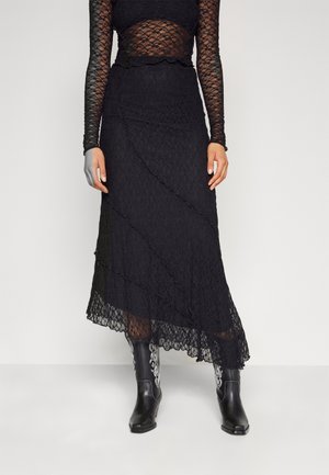 Vrouw die een zwarte kanten top met lange mouwen draagt en een bijpassende kanten midi-rok met zwarte geborduurde cowboylaarzen, staand tegen een effen achtergrond.