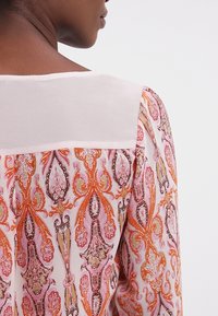 Långärmad blus med ett intrikat paisley-mönster i orange, rosa och brunt; slät vit ståndkrage på baksidan; lättviktig tyg.