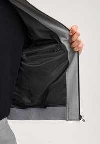 Grau Jacke mit schwarzem Futter, ausgestattet mit einer Reißverschlusstasche innen und einer leicht strukturierten Außenseite. Das Innere ist glatt und elegant.