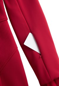 Piros szövetkabát bordázott textúrával az ujján, rejtett zsebbel, amelyen fehér címke látható. Egységes, sima felület.