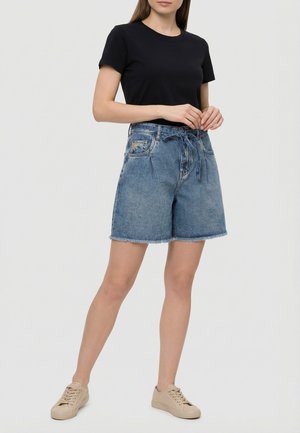 Vrouw die een zwart T-shirt draagt, een hoge taille blauwe spijkershorts met een voorstrik en beige sneakers, staand tegen een effen achtergrond.