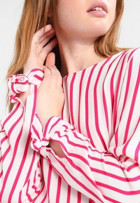 Bovenlichaam van een vrouw in een witte blouse met verticale roze strepen, handen die haar nek en haar aanraken, en mouwen die in knopen op de polsen zijn gestrikt.