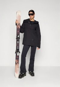 Schwarze Skijacke mit Reißverschluss, kombiniert mit eng anliegenden schwarzen Skihosen. Die Person hält einen Ski mit Bindungen und trägt große schwarze Sonnenbrillen.