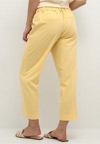 Pantalons jaunes cropped avec une taille élastique, deux poches arrière, texture lisse et une finition soignée à la cheville. Associés à des sandales beiges.