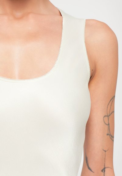 Närbild av övre delen av kroppen som bär en ärmlös krämfärgad topp med en minimalistisk linjeteckningstatuering på höger arm.