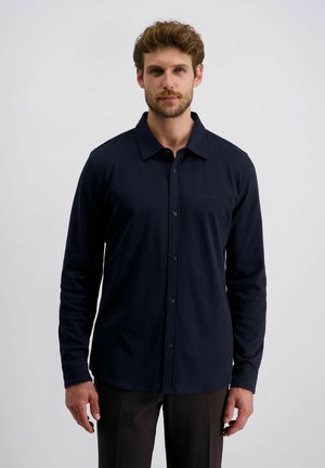 Homme aux cheveux courts et bouclés avec une barbe, portant une chemise en jean à manches longues de couleur navy et un pantalon sombre, se tenant devant un fond clair.