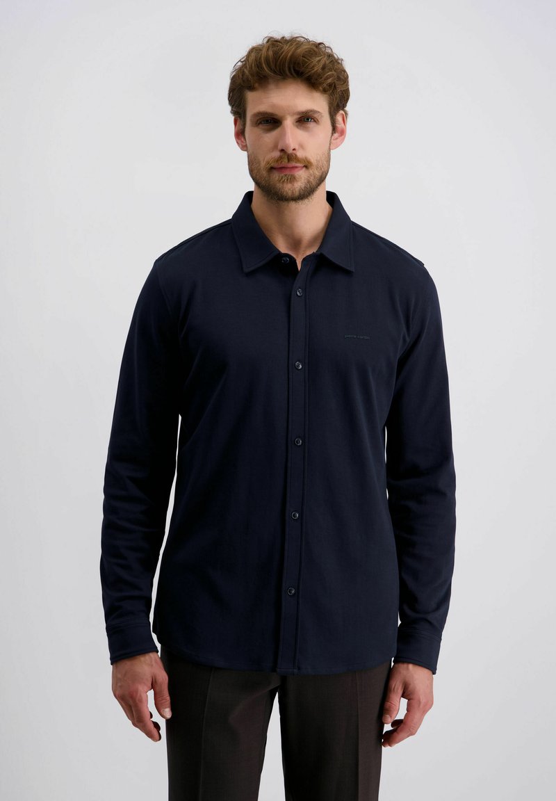 Man met kort krullend haar en baard, gekleed in een marineblauwe blouse met lange mouwen en donkere broek, staand tegen een lichte achtergrond.