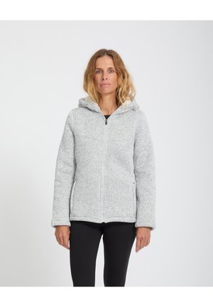 Donna che indossa una giacca con cappuccio grigio chiaro con zip e pantaloni neri, in piedi davanti a uno sfondo bianco uniforme.