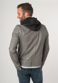 Grijze leren jas met een afneembare zwarte capuchon, met een gestroomlijnd ontwerp en zichtbare stiksels. Draagt over spijkerbroeken.