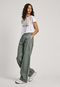 Svijetlosiva majica s šarenim tekstom "Pepe Jeans", u kombinaciji s širokim maslinasto-zelenim cargo hlačama koje imaju bočne džepove. Bijeli i sivi tenisice.