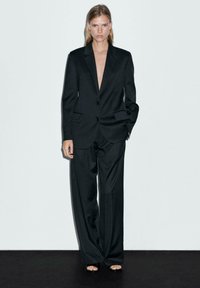 Black suit with a deep V-neckline, two buttons, and wide-leg trousers. Features large pockets and minimalistic design, paired with open-toe shoes.
