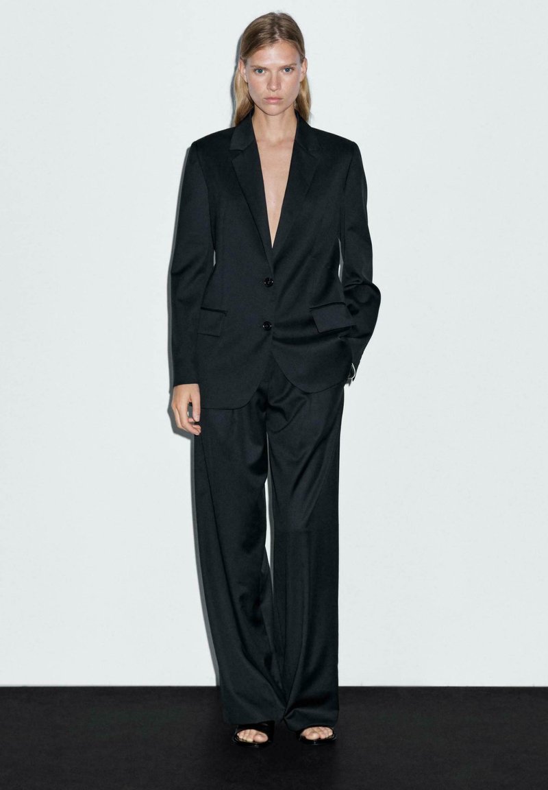 Zwarte suit met een diepe V-hals, twee knopen en wijde broekspijpen. Bevat grote zakken en een minimalistisch ontwerp, gecombineerd met open-sandalen.