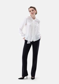 Blusa bianca con collo a fiocco e maniche lunghe, abbinata a pantaloni neri su misura e tacchi a punta neri. Tessuto dalla texture liscia.
