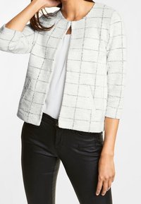 Vit texturerad jacka med ett rutmönster, rund halsringning och trekvartsärmar, kombinerad med svarta byxor.