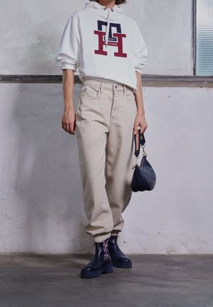 Person trägt weißen Tommy Hilfiger Hoodie, beige Hose, schwarze Stiefel mit lila Socken, hält eine kleine schwarze Tasche, steht drinnen.