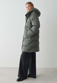 Doudoune vert olive avec capuche, dotée d'une fermeture éclair frontale, de poches latérales et d'une coupe légèrement ample. Portée sur un pantalon large noir.