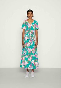 Mulher a usar um vestido envolvente floral em verde-água com mangas curtas e ténis brancos, de pé contra um fundo claro e simples.