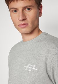 Giovane uomo con capelli corti che indossa una felpa grigia chiara con testo bianco "LES DEUX COPENHAGEN EST. 2011" sul petto.