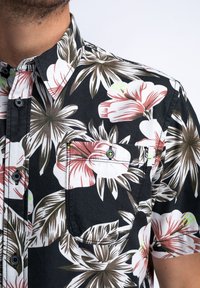 Camisa de manga corta con estampado floral en negro, que presenta patrones de hibiscos blancos y rosas, y un bolsillo en el pecho con un detalle en verde de contraste.