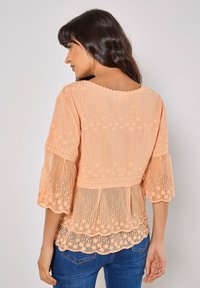 Pfirsichfarbene Bluse mit strukturierten Stickereien, glockigen Ärmeln und einem gewellten Saum. Transparenter Stoff mit floralen Mustern, schichtendes Design.