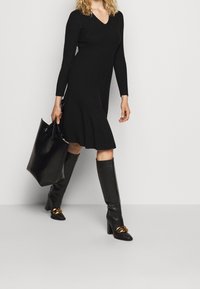Abito nero a coste con scollatura a V, abbinato a stivali al ginocchio in pelle nera con dettagli in catena dorata e una borsa tote nera.