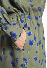 Olivově zelené šaty s modrým květinovým vzorem a zelenými akcenty, s elastickým pasem, knoflíkovými manžetami a bočními kapsami. Měkká textilie.