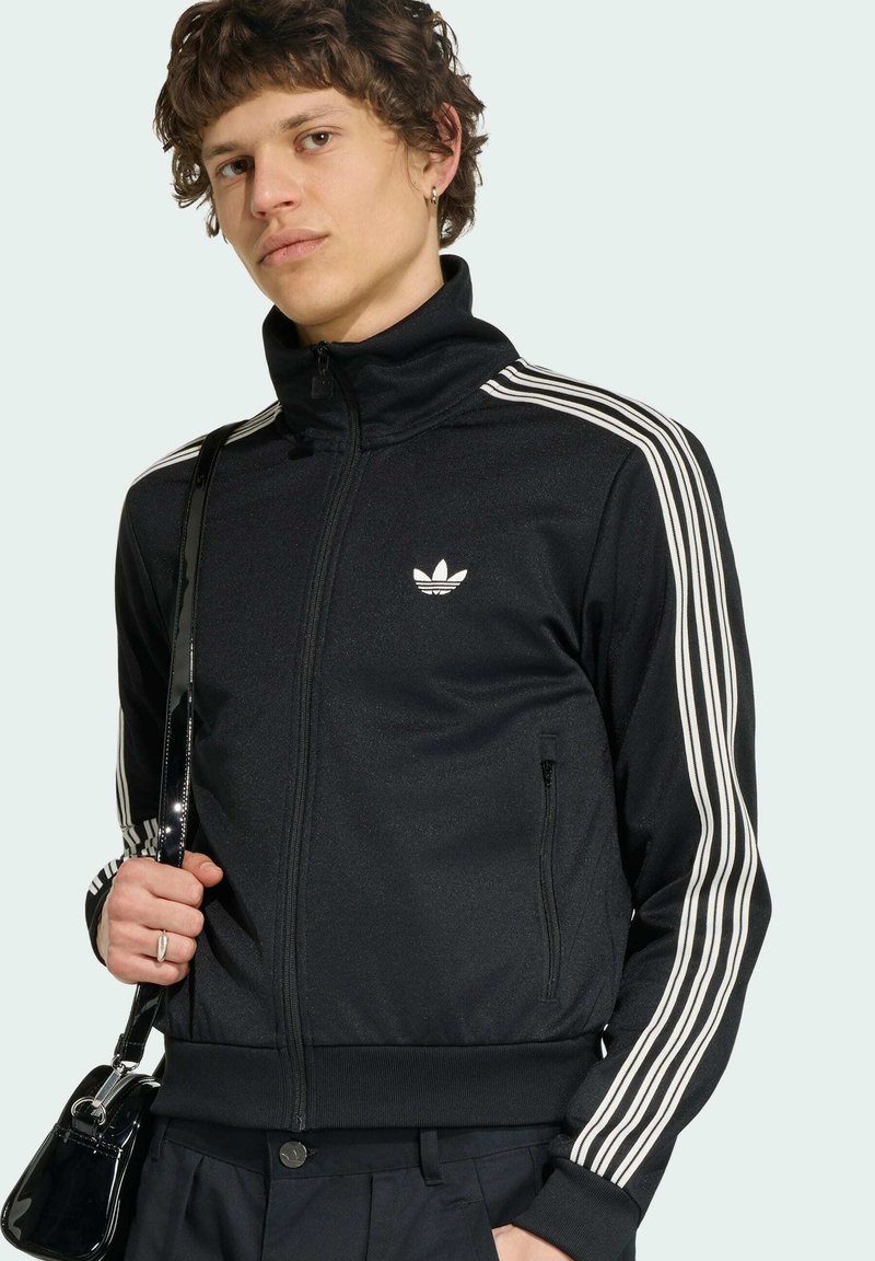Junger Mann mit lockigem Haar, trägt eine schwarze Adidas Zip-Jacke mit weißen Streifen und hält eine schwarze Handtasche über einer Schulter.