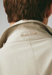 Widok z tyłu osoby w beżowym płaszczu z wyszytym napisem "Massimo Dutti" na kołnierzu, pod krótkimi brązowymi włosami.