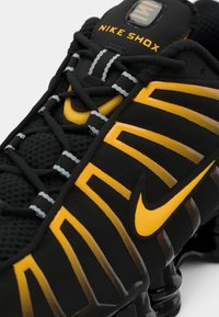 Nike Shox apavi ar melnu sietu augšējo daļu, dzelteniem akcents un izteiksmīgu svītru dizainu. Iekļauj strukturētu virsmu un mīkstu zoli.