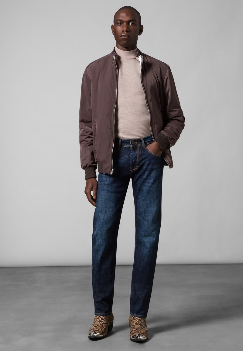 Dark brown bomber jacket, beige turtleneck, blue jeans, and brown snake-print shoes. Model stands against a neutral gray background.